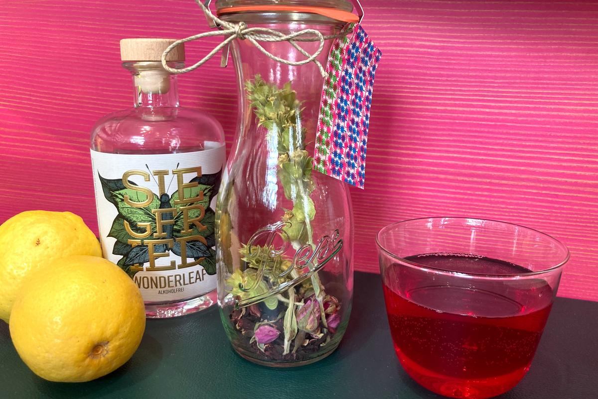 Zwei Zitronen, alkoholfreier Gin von SIEGFRIED und MocktailMischung in der WECK-Flasche mit Hibiskus, Rose und Bergtee. Rechts daneben steht ein Glas mit dem Mocktail in roter Farbe.