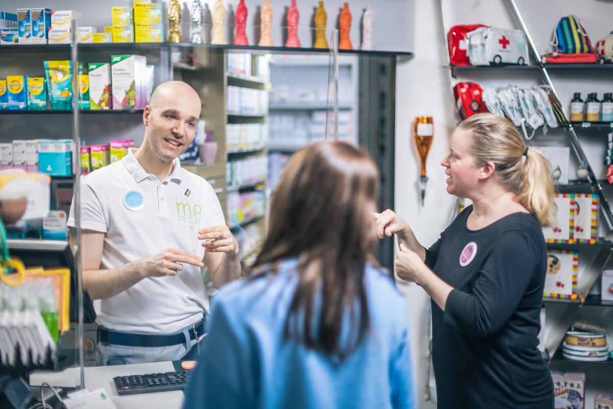Mag. Sreco Dolanc an der Tara in der Marien Apotheke Wien. Er hat ein Beratungsgespräch mit einer Kundin, seine Gebärdensprachdolmetscherin steht rechts neben ihm. Die Kundin ist nur von hinten sichtbar.