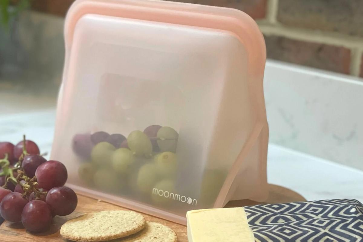 Silikonbeutel in rosa auf Holzbrett mit Weintrauben, Keksen und Käse