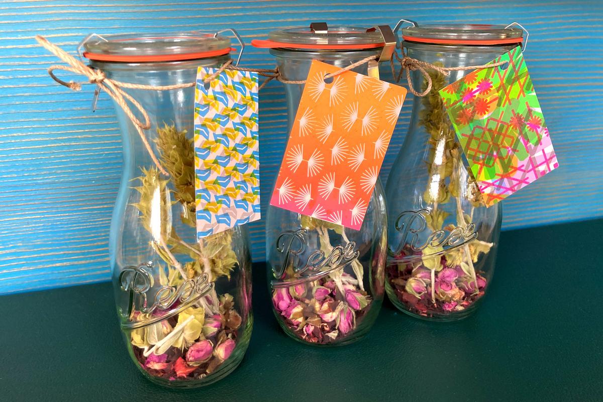 Drei WECK-Flaschen mit Hibiskusblüten, Rosenknospen und Rispen von Griechischem Bio-Bergtee, die auf einer Tara-Ablage in der Marienapotheke Wien stehen