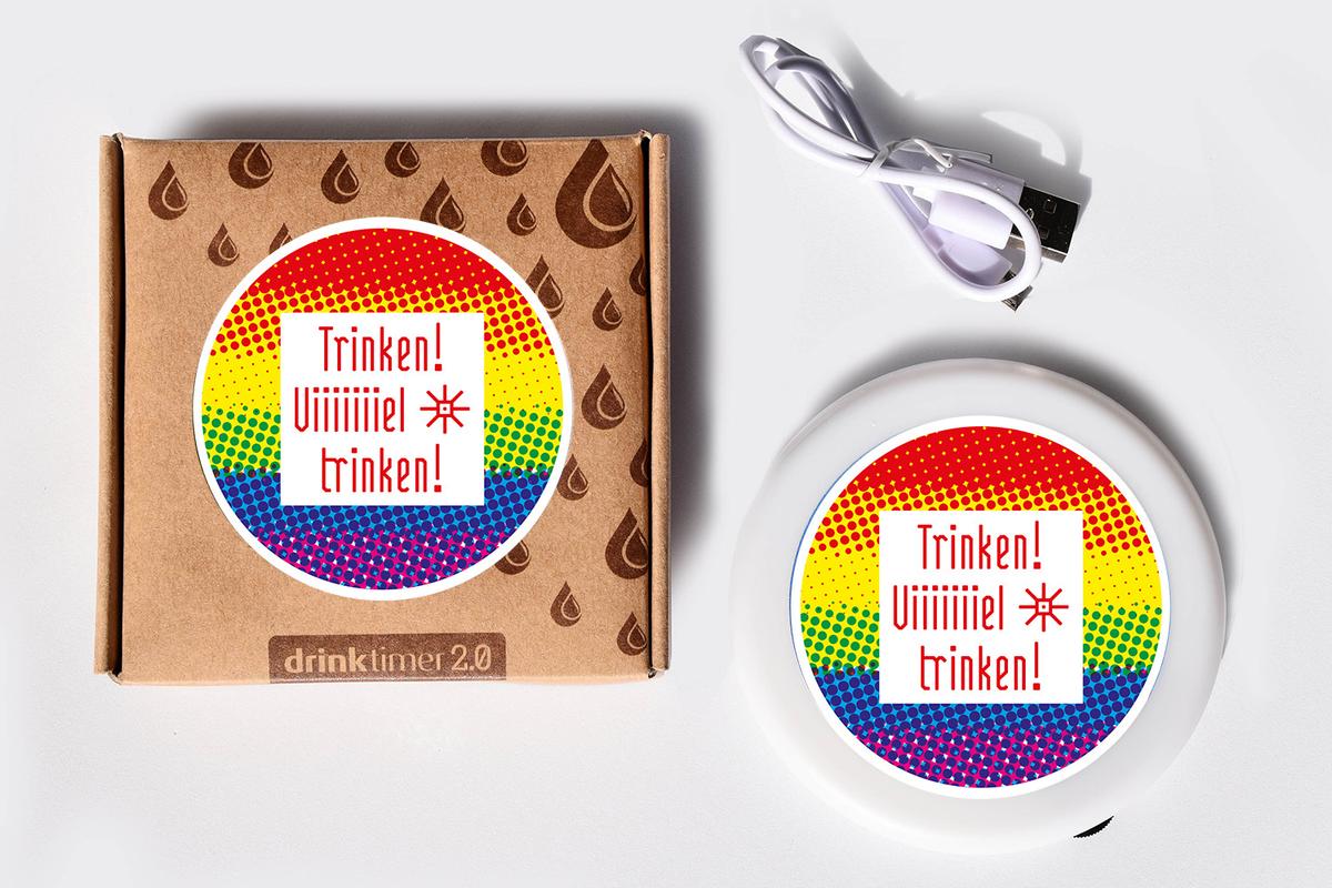 Der intelligente Untersetzer mit Trinkerinnerung, der Drink Timer, in einer Schachtel und daneben offen mit Ladekabel. Der Drink Timer ist von der Marien Apotheke Wien individualisiert und der Spruch auf dem regenbogenbunten Sticker lautet "Trinken. Viiiiiiiiiiiel trinken!"