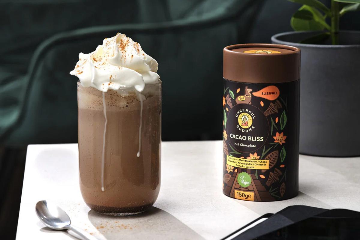 Cacao Bliss Mischung von Cheerful Buddha. Links steht ein großes Glas Kakao mit Schlagobers-Haube, daneben liegt ein Löffel. Rechts davon steht die Packung mit der Kakao-Mischung "Cacao Bliss" von Cheerful Buddha mit Vitalpilzen.