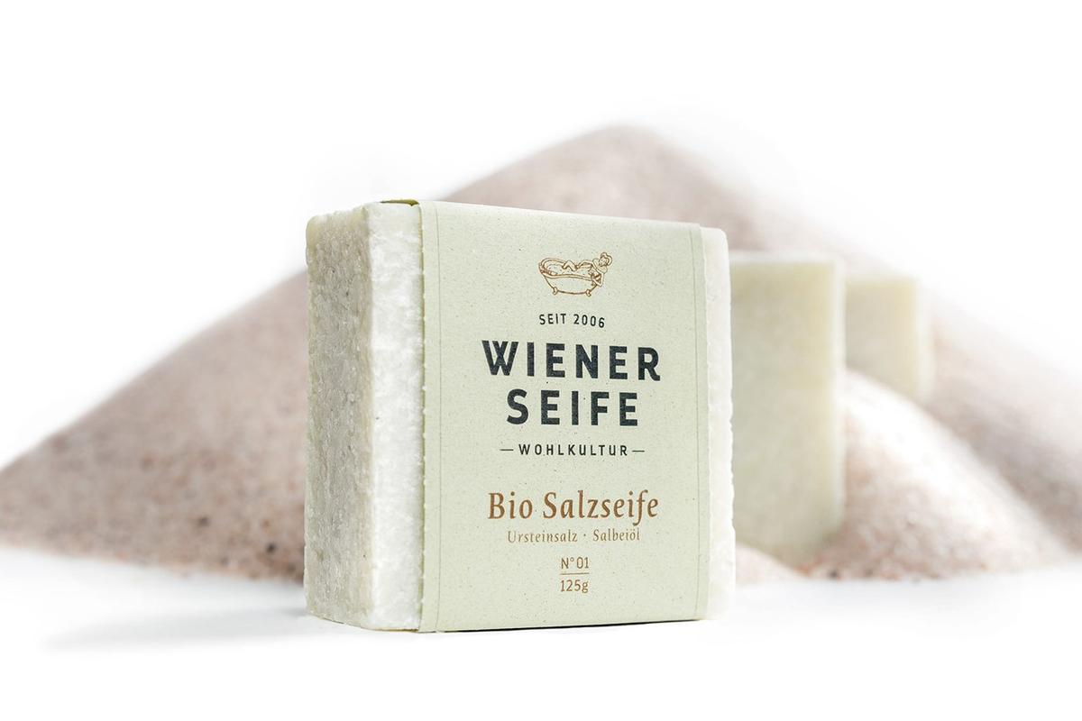 Weißes Seifenstück verpackt in Papierumschlag mit der Aufschrift "Wiener Seife" - Bio Salzseife. Im Hintergrund befindet sich eine Menga an aufgeschüttetem Salz.