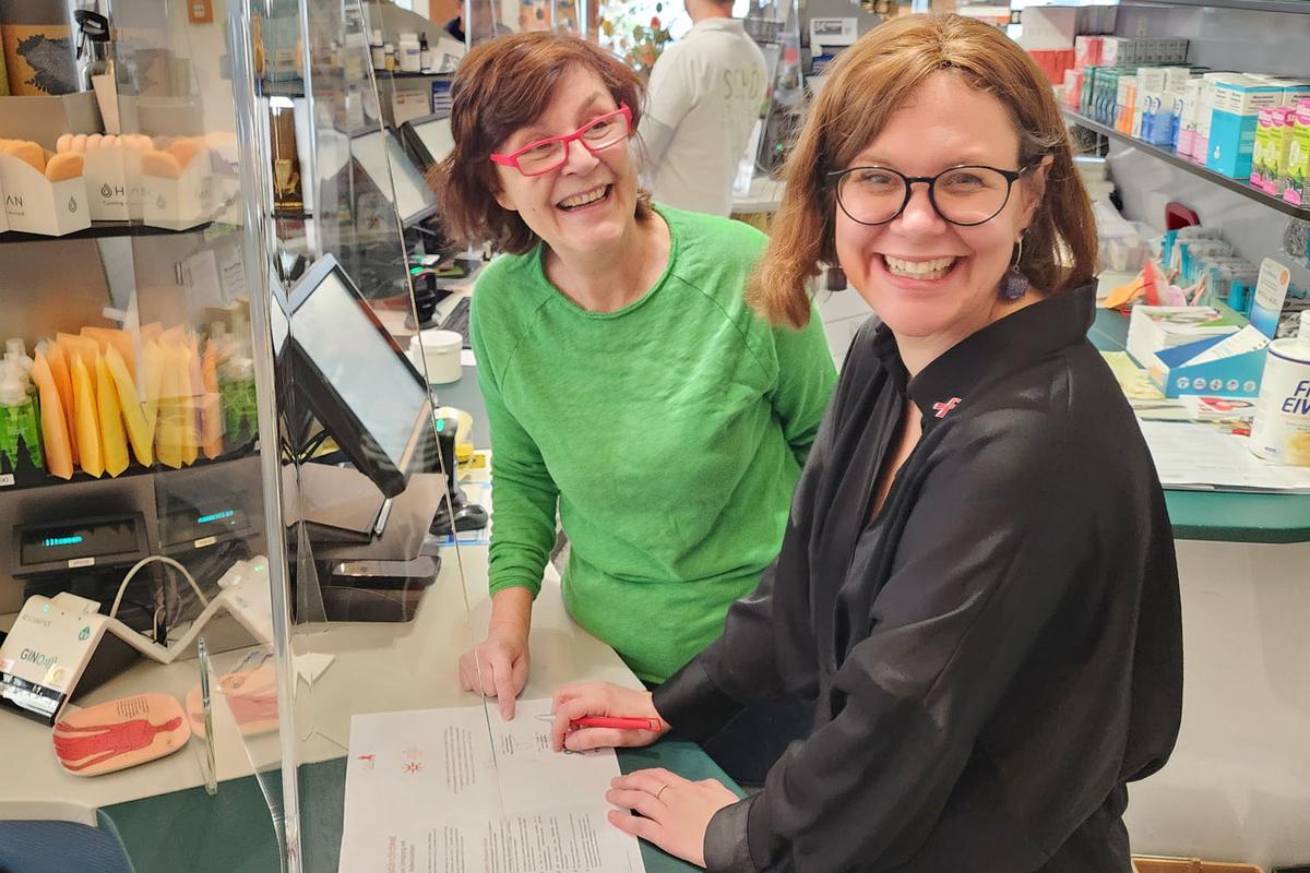 Mag. pharm. Karin Simonitsch und Andrea Brunner, Geschäftsführerin der Aids Hilfe Wien, stehen in der Marien Apotheke Wien hinter der Tara und unterzeichnen die Unterstützungserklärung zu #positivarbeiten.