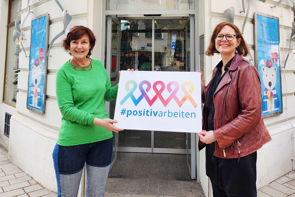 Mag. Karin Simonitsch und Andrea Brunner, Geschäftsführerin der Aids Hilfe Wien, stehen vor der Marien Apotheke in 1060 Wien. Sie halten gemeinsam ein Schild mit der Aufschrift "positivarbeiten" und vielen bunten Ribbons.