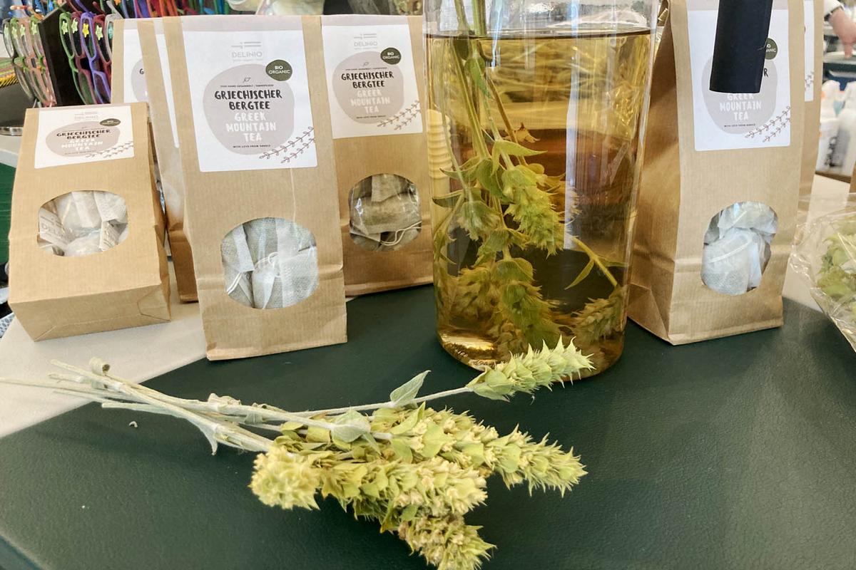 Mehrere Packungen mit Teebeuteln von Griechischem Bio-Bergtee, einer Bergtee-Rispe im Vordergrund und einer Teekanne mit Rispe im Hintergrund